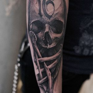 image of an arm tattoo featuring a realistic human skull and crossbones with the number 16 overlayed onto the skull forehead as meaning for the client