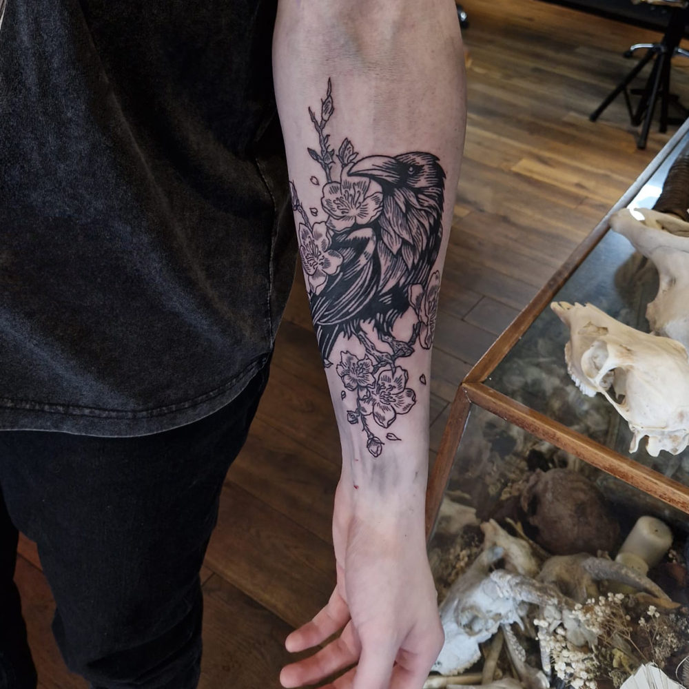 luke-tattoo-crow-cherry-blossoms photo of an arm tattoo showing a crow with cherry blossoms and branches in a blackwork etching style