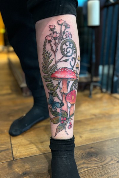image of a leg tattoo which features mushrooms, foxglove flowers, twirling leaves and vines all done in bright colours.