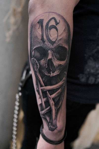 image of an arm tattoo featuring a realistic human skull and crossbones with the number 16 overlayed onto the skull forehead as meaning for the client